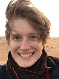 Headshot of smiling woman Alex Thomson with windblown, short dark blond hair in sandy desert background