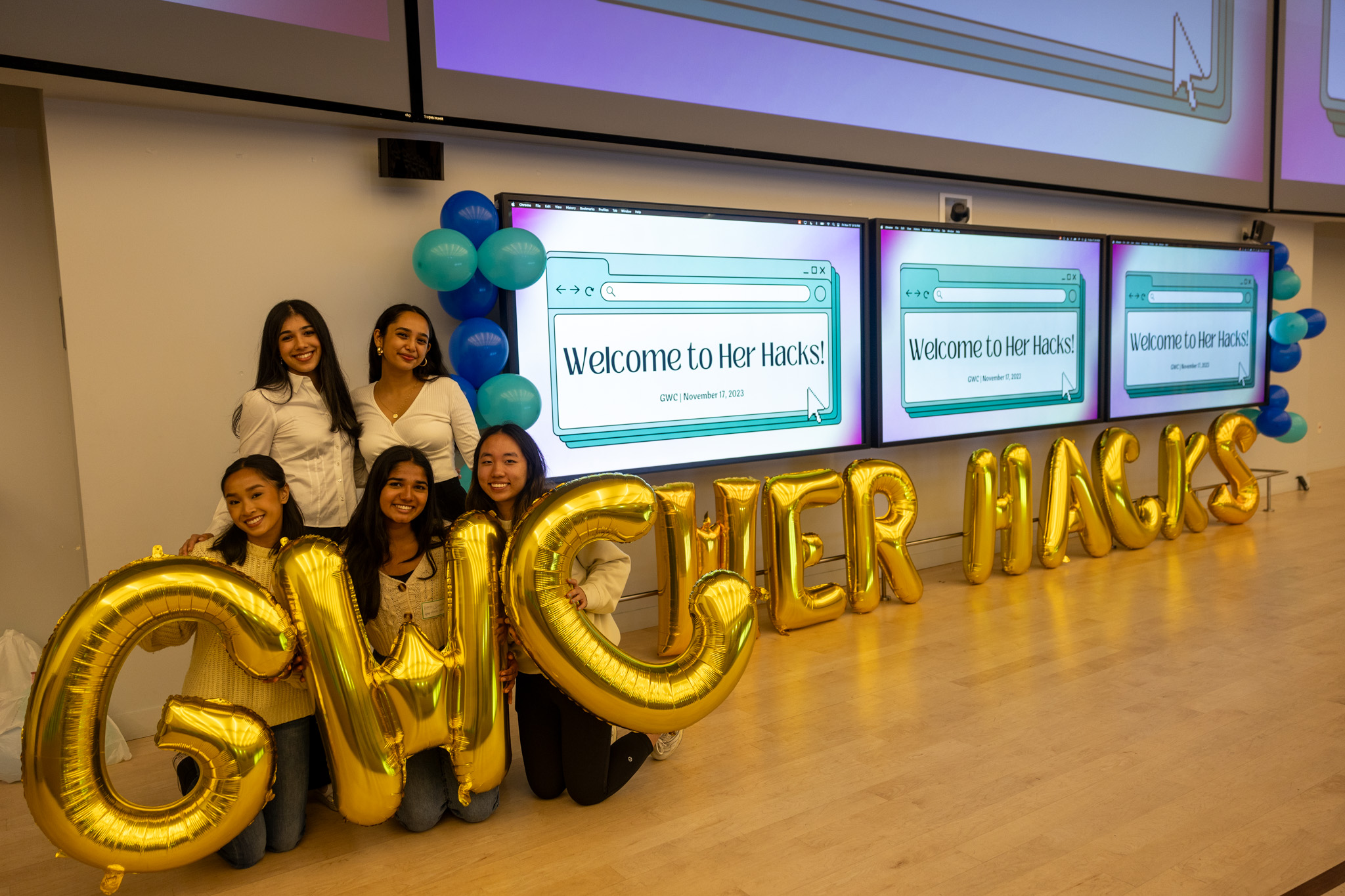 Two girls are standing and three girls are kneeling on stage next to screens that read Welcome to Her Hacks