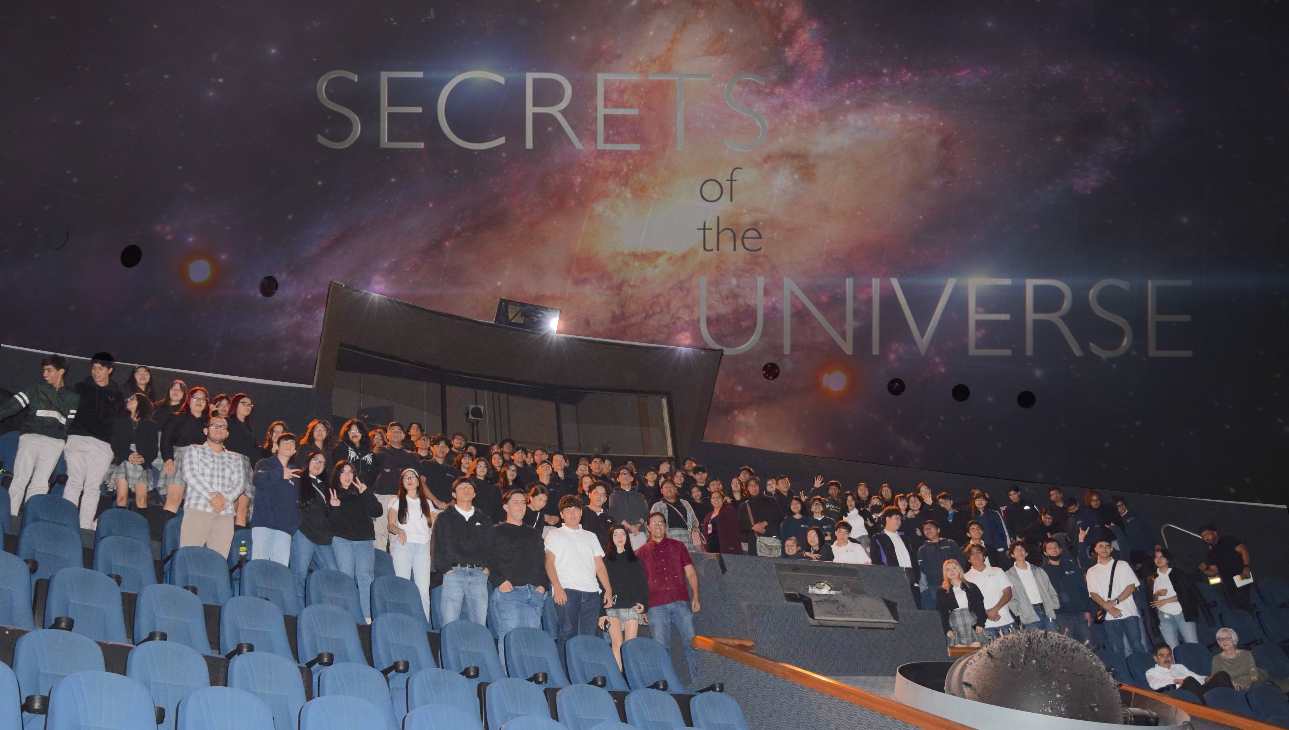 Manuel Calderón de la Barca Sánchez is standing with a group of students in a large theater with the words Secrets of the Universe written in the background 
