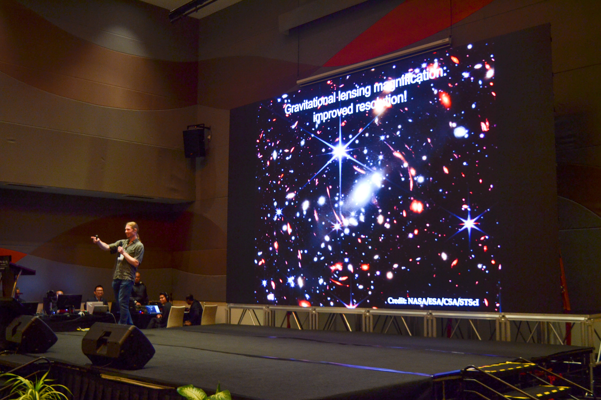 Tucker Jones stands center stage while delivering a talk. Behind him, a screen shows an image of the universe. 