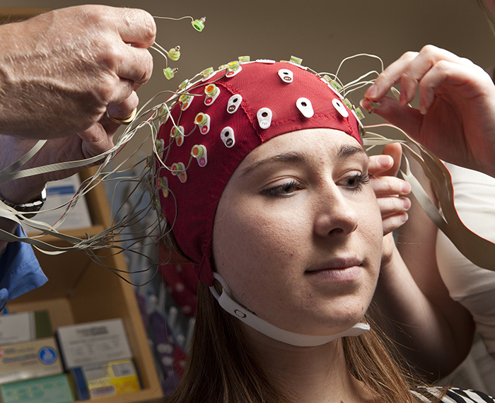hands of two people attaching electrodes to a red cap atop the head of a young woman.