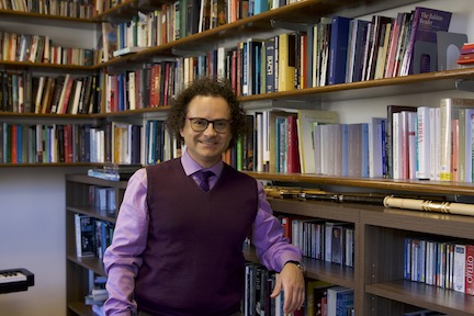 man in purple shirt and sweater vest, wearing glasses, leaning on bookshelf