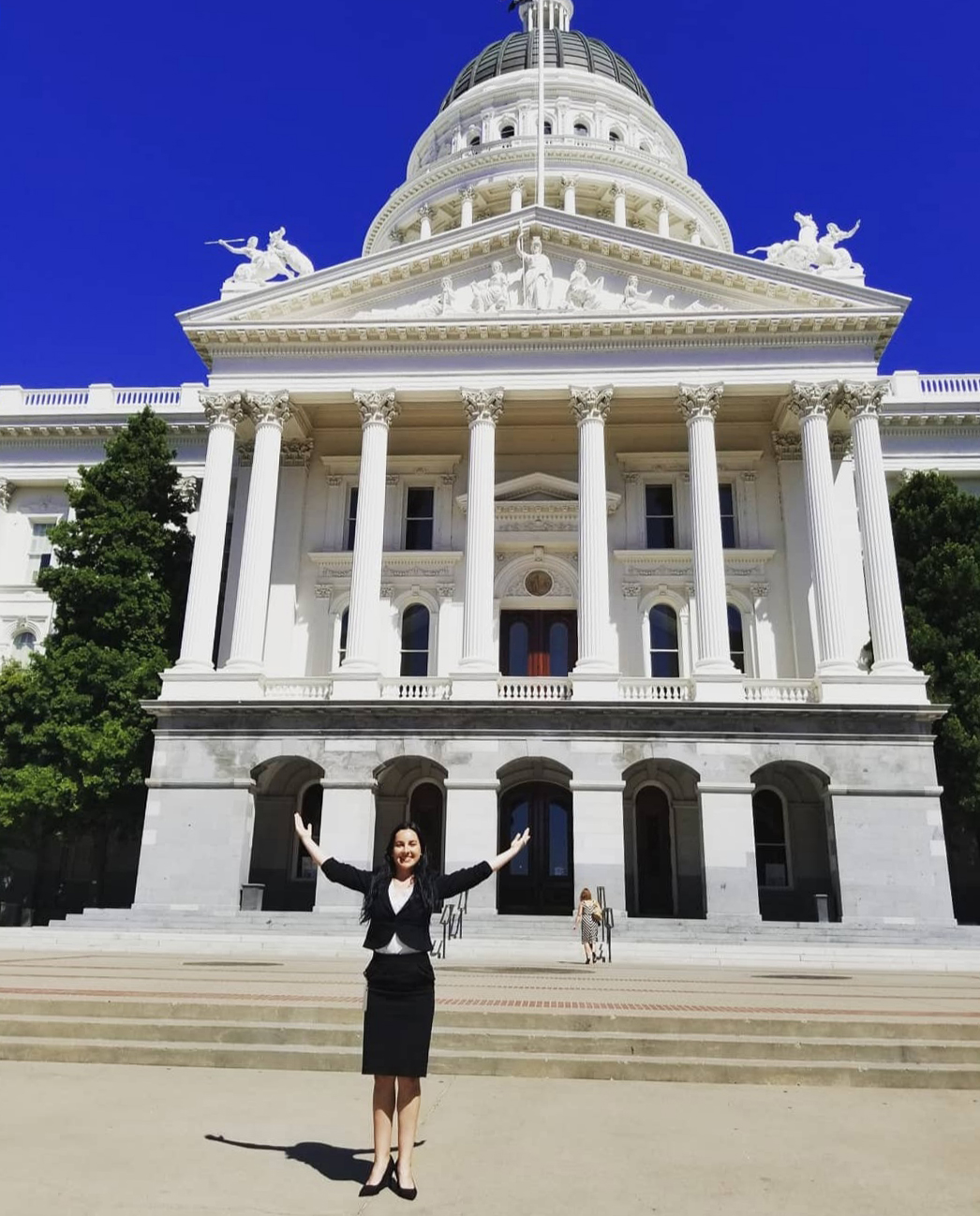 young woman in business suit standing in front of California capitol building in Sacramento with arms upraised