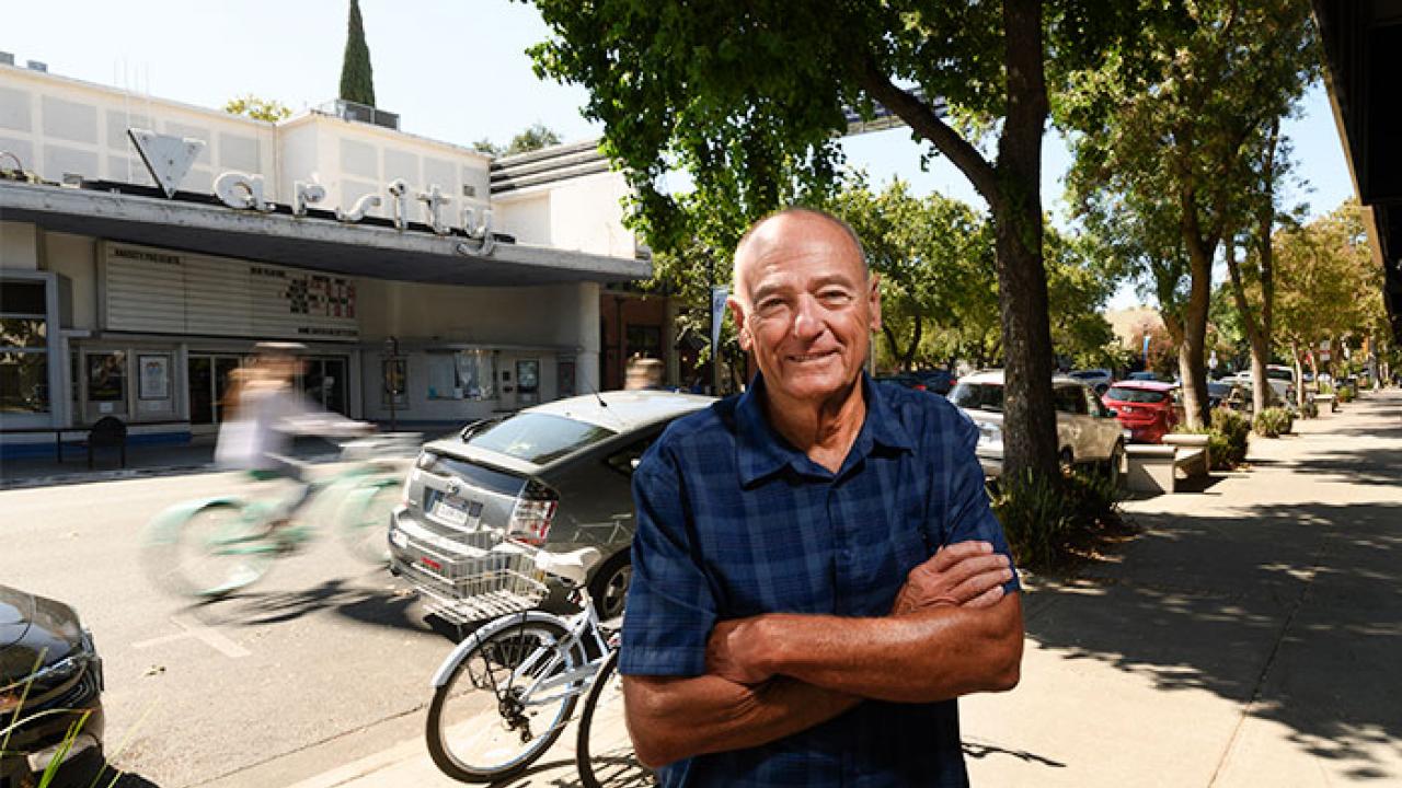 Photo of Bob Dunning with arms folded, standing on downtown Davis sidewalk, Varsity Theater, bikes and parked cars in background.