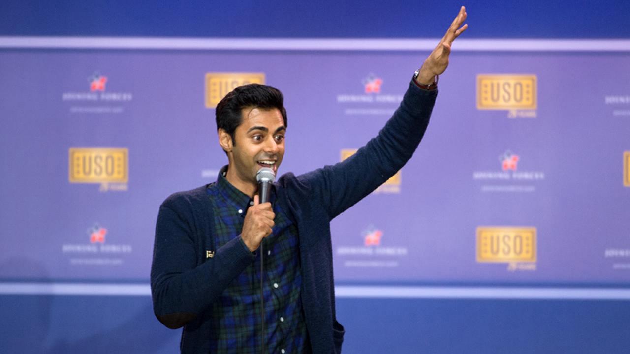 Photo: Hasan Minhaj gesturing with a raised hand during a show. 