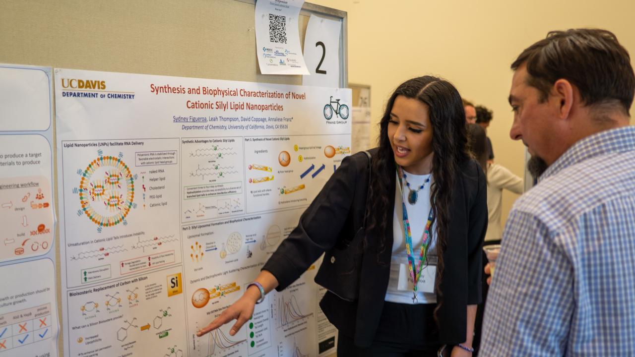 A student points to a poster as a bystander listens to her talk about research
