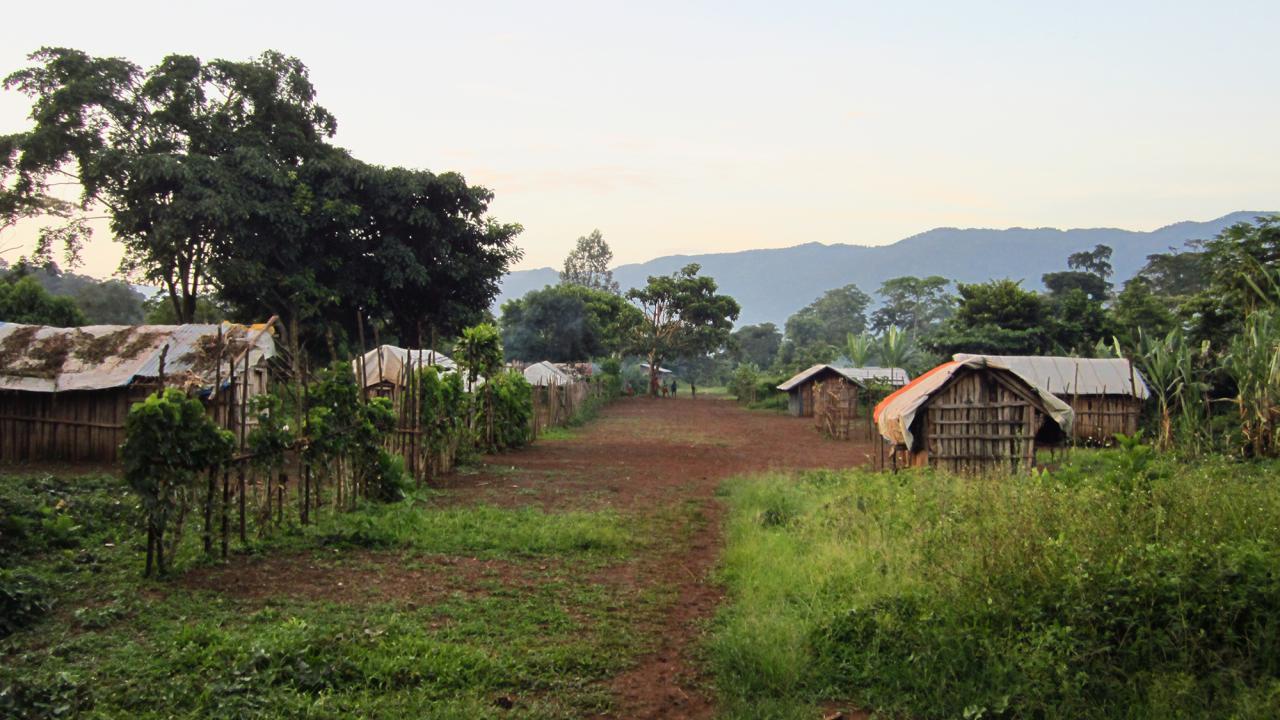Looking down the middle of an African village with wooden buildings, a few people in the distance and  and mountains farther off.