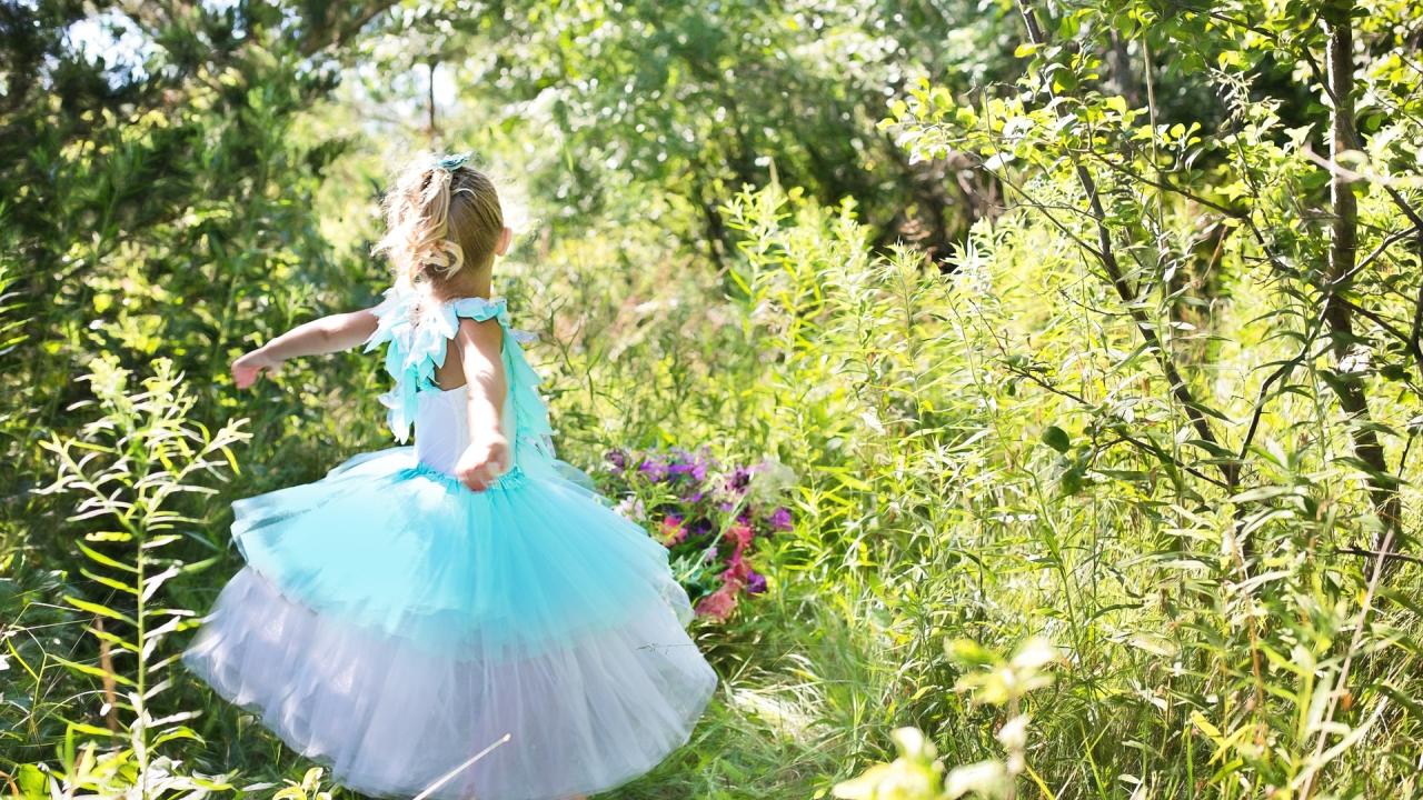 A little girl with a blond ponytail and a blue and white dress dances around a wooded area with flowers