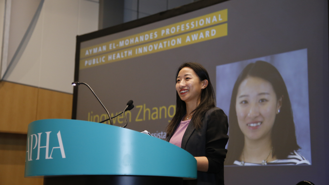UC Davis communication faculty Jingwen Zhang standing at podium with APHA written on front. Behind her a large screen shows her name, photo and the words “Ayman El-Mohandes Young Professional Public Health Innovation Award.”