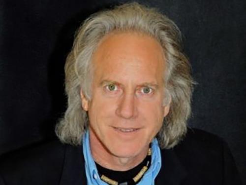 Headshot of older man with long grey hair and exposed forehead, blue eyes, blue collar shirt with black coat