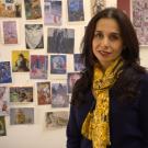 Shiva Ahmadi is wearing a colorful yellow scarf and navy sweater and sitting in front of a wall pull of different pictures